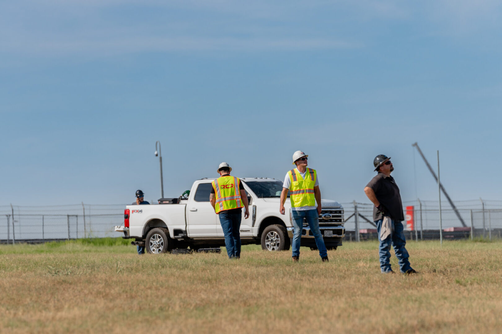 Two construction workers and a man in casual clothes stand near a white pickup truck on a grassy field.