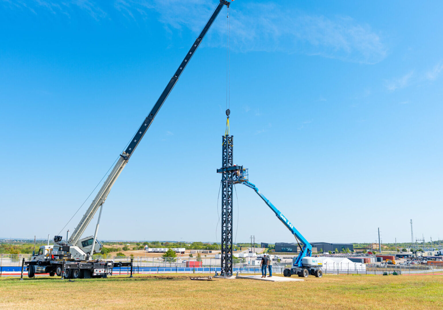 Construction workers lifting a tall steel structure using cranes under a clear blue sky.