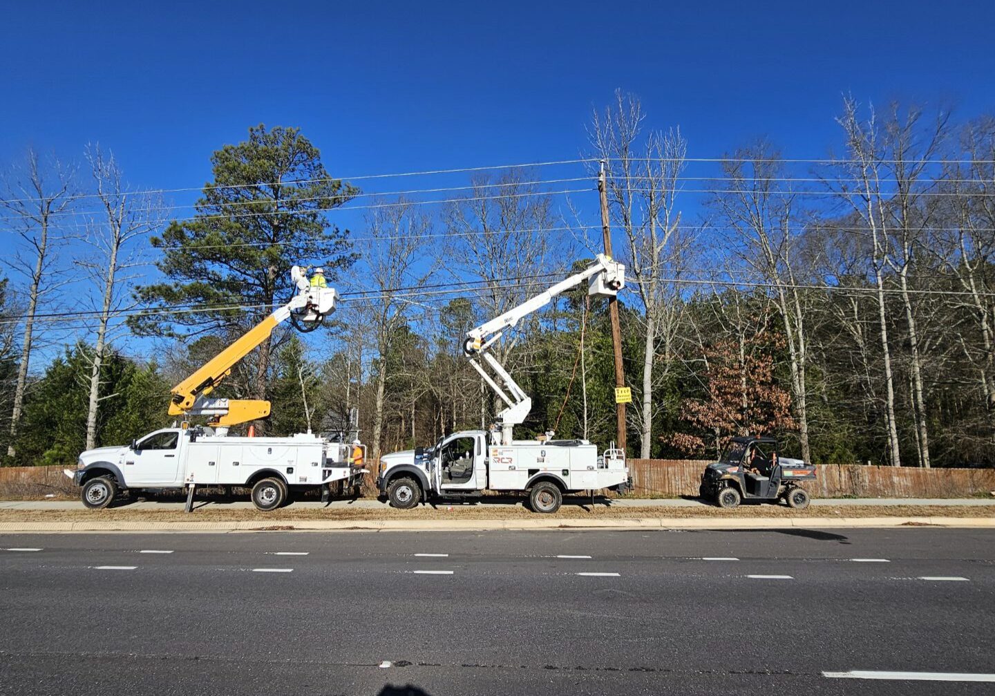 Two utility trucks with extended booms working on power lines by the roadside.